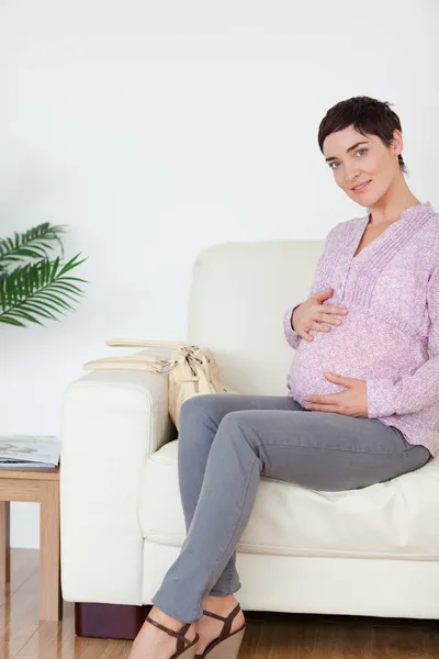 Mulher muito grávida sentada em um sofá tocando sua barriga — Fotografia de Stock