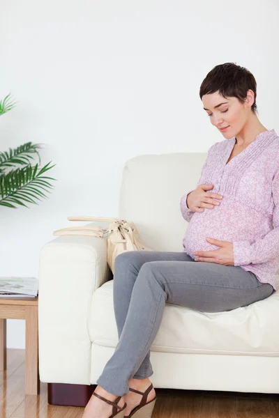 Femme enceinte souriante assise sur un canapé touchant son ventre — Photo