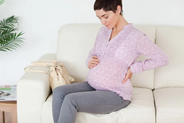 魅力的な妊娠中の女性に触れる彼女の腹のソファーに座っています。 — ストック写真