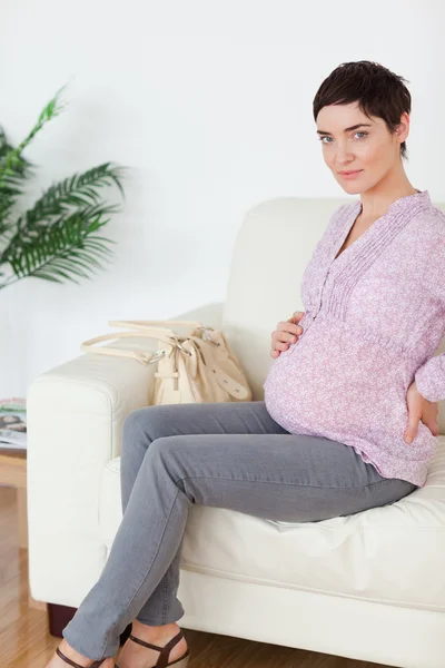 Brünette schwangere Frau sitzt auf einem Sofa und berührt ihren Bauch — Stockfoto