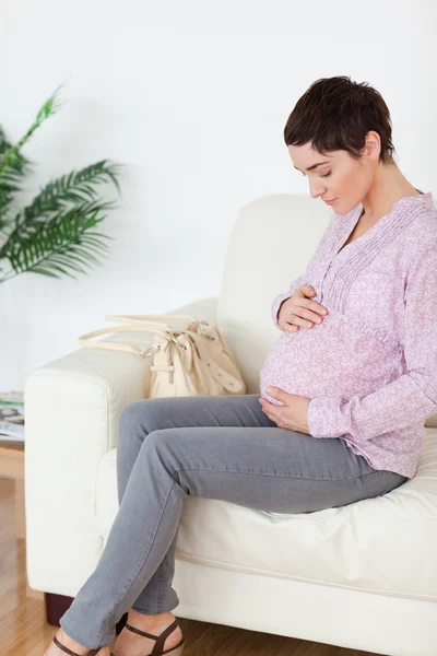 Brunette femme enceinte aux cheveux courts assise sur un canapé touchant — Photo