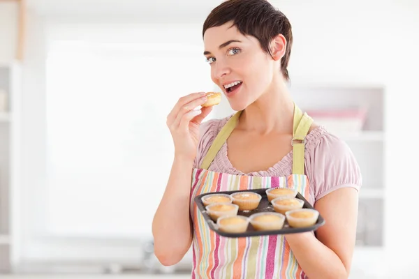 Mulher morena alegre mostrando muffins enquanto come um — Fotografia de Stock