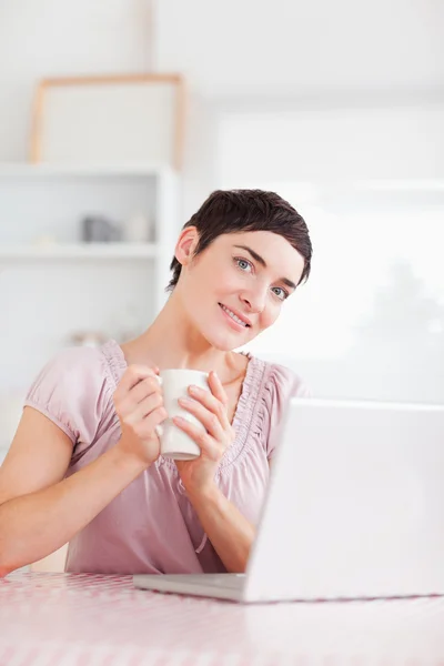Cute woman with a cup and a laptop smiling into the camera — Stock Photo, Image
