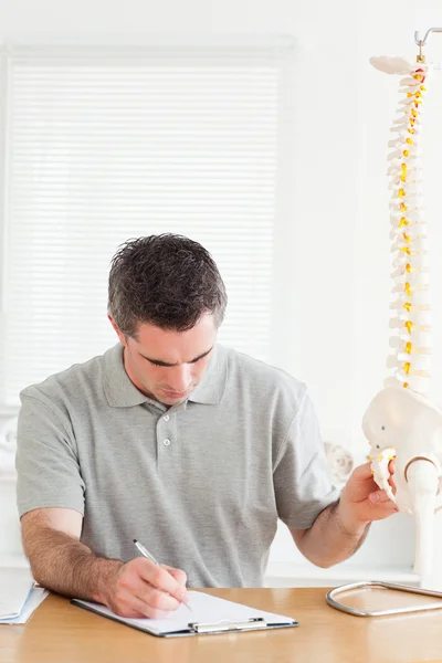 Doctor macho sentado en una mesa trabajando con una columna vertebral modelo — Foto de Stock