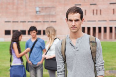 Lonely male student posing while his classmates are talking clipart