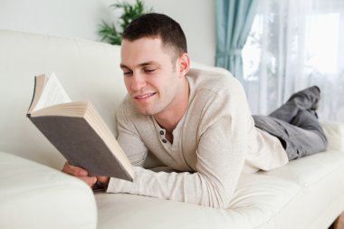 Young man lying on a couch to read a book clipart