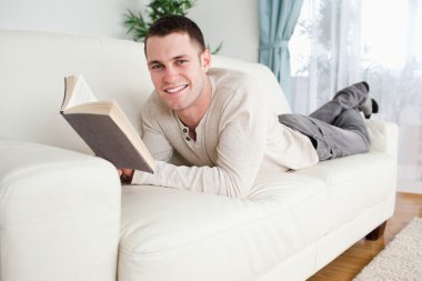 Young man lying on a couch reading a book clipart