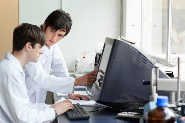 Chemist pointing at something on a monitor to his student — Stock Photo, Image