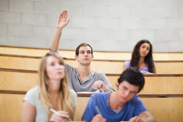 Estudiante levantando la mano mientras sus compañeros de clase están tomando notas —  Fotos de Stock