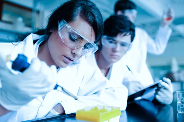 Jovem cientista deixando cair líquido em tubos de ensaio, enquanto seu parceiro está tomando notas — Fotografia de Stock