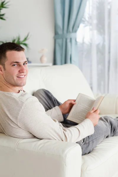 一个微笑的人拿一本书坐在沙发上躺的肖像 — 图库照片