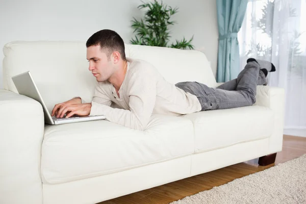 Homem bonito deitado em um sofá usando um laptop — Fotografia de Stock