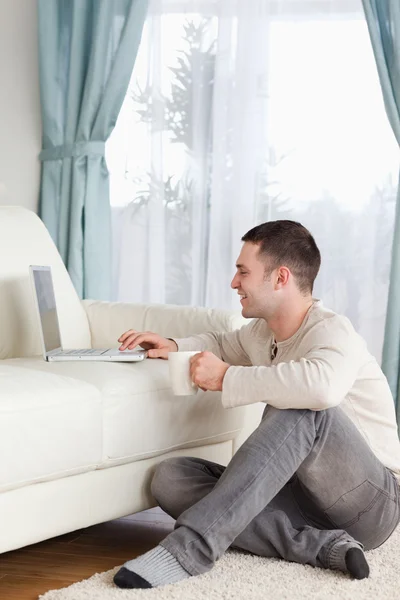 Πορτρέτο ενός άντρα που κάθεται σε ένα χαλί, χρησιμοποιώντας ένα φορητό υπολογιστή και κρατώντας — Φωτογραφία Αρχείου