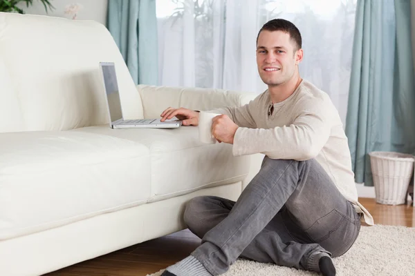 Mann sitzt mit einer Tasse Tee auf einem Teppich und benutzt einen Laptop — Stockfoto