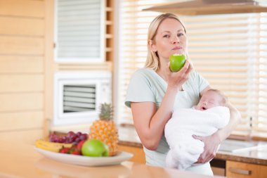 Woman chewing on apple while holding baby on her arms clipart