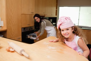 Girl sitting in the kitchen while her mother puts cookies into t clipart