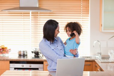 Mother and daughter using notebook and cellphone in the kitchen clipart