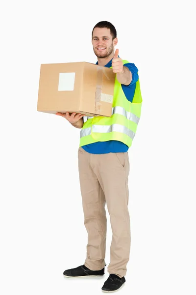Sonriente joven repartidor dando el pulgar hacia arriba mientras sostiene el paquete —  Fotos de Stock