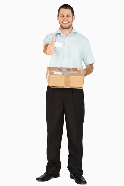 Smiling young post employee with parcel giving thumb up — Stock Photo, Image