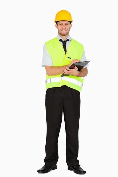 Smiling young foreman in safety jacket taking notes on his clipb — Stock Photo, Image