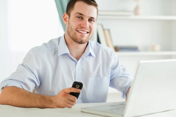 Biznesmen uśmiechający się trzymając jego komórki podczas pisania na swoim laptopie — Zdjęcie stockowe