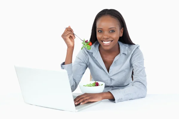 Joyful businesswoman working with a laptop while eating a salad — Stock Photo, Image