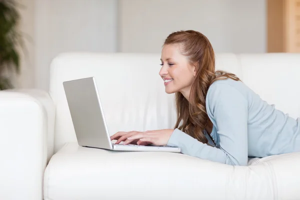 Veselá usmívající se žena na pohovce, psaní na její laptop — Stock fotografie