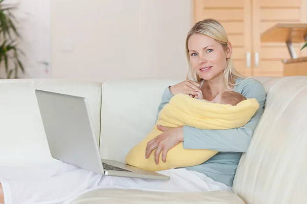 彼女は赤ん坊を保持しているソファの上のラップトップを持つ母 — ストック写真