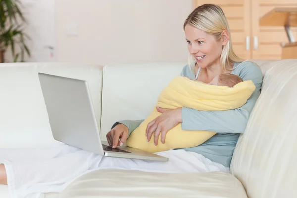 Matka z laptopem na kanapie, trzymając jej dziecko — Zdjęcie stockowe
