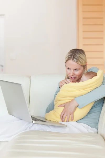 Mother with notebook on the couch holding her baby — Stock Photo, Image