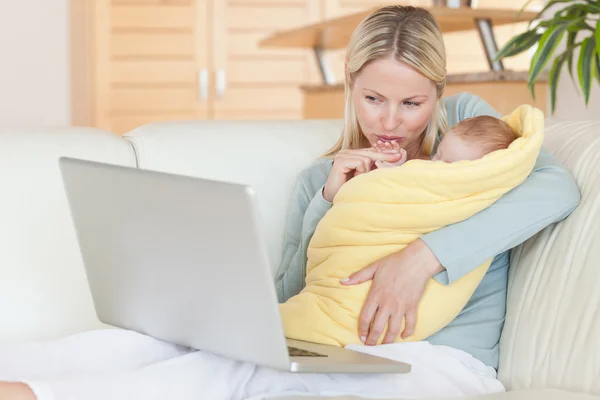 Mother holding her baby while looking at her laptop — Stock Photo, Image