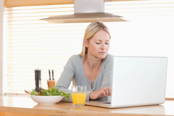 Mulher com salada e laranja trabalhando no laptop na cozinha — Fotografia de Stock
