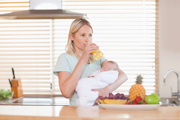 Woman taking a sip of juice while holding her baby — Stock Photo, Image