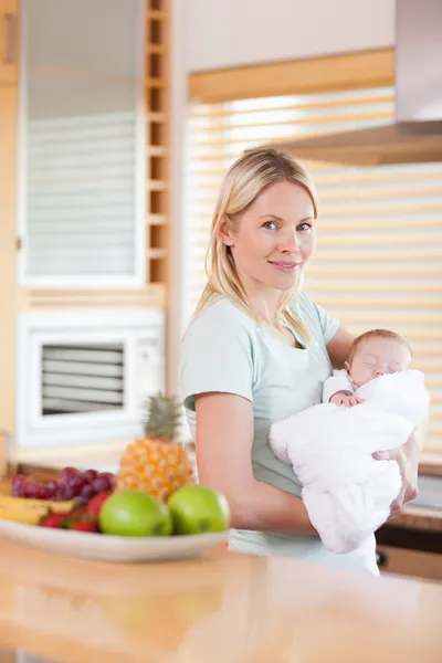 Vista lateral de la mujer de pie en la cocina con su bebé en su — Foto de Stock