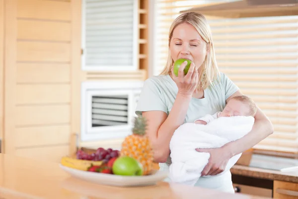 Woman eating an apple while holding her baby — Stock Photo, Image