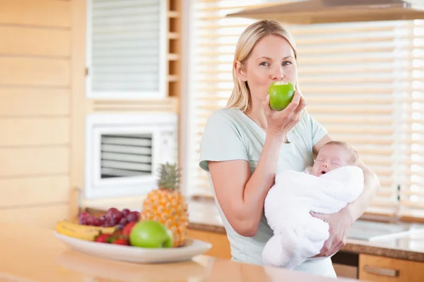 Woman enjoying an apple with her baby on her arms — Stock Photo, Image