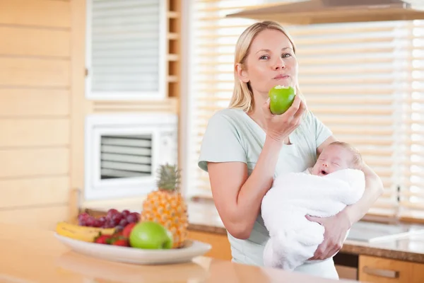 Woman chewing on apple while holding baby on her arms — Stock Photo, Image