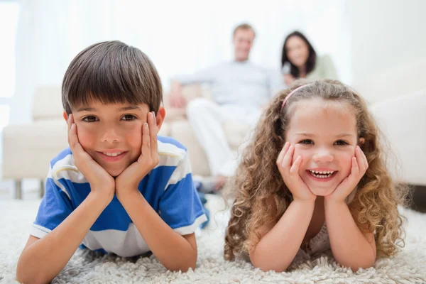 Szczęśliwe dzieci leżąc na dywanie z rodzicami za nimi — Zdjęcie stockowe