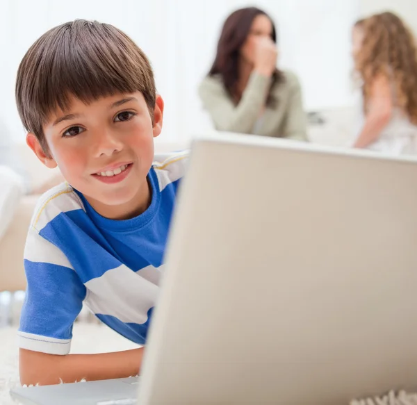 Menino usando laptop no tapete com sua família atrás dele — Fotografia de Stock