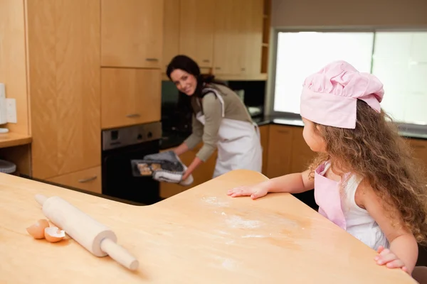 Girl watching her mother putting cookies into the oven — Stock Photo, Image