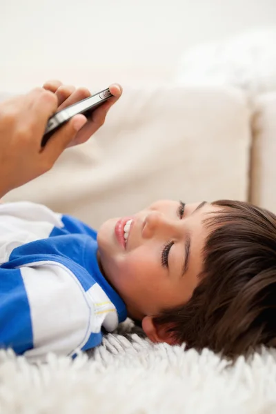 Jongen speelt met zijn cellphone liggend op het tapijt — Stockfoto