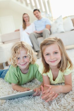 Siblings on the carpet with tablet and parents behind them clipart