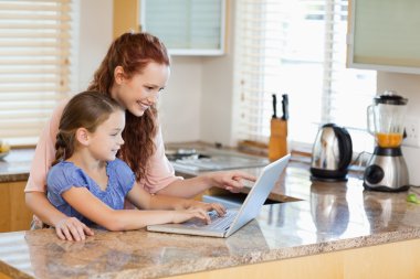 Mother and daughter with laptop behind the kitchen counter clipart