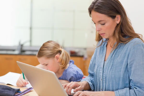 Liten flicka gör sina läxor medan hennes mamma använder laptop — Stockfoto