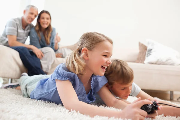 Siblings playing video games with their parents on the backgroun — Stock Photo, Image