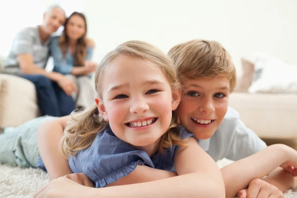 Happy siblings posing on a carpet with their parents on the back — Stock Photo, Image