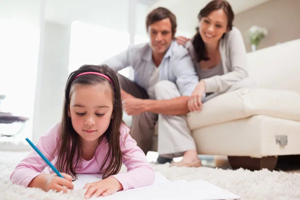 Little girl drawing with her parents in the background — Stock Photo, Image