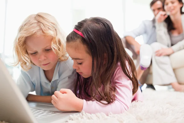 Syskon som använder en bärbar dator medan deras föräldrar är i backgro — Stockfoto