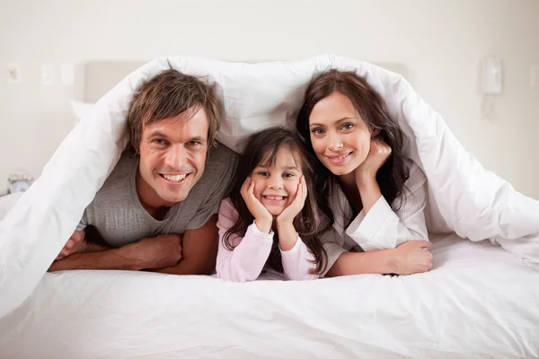 Sorrindo pais deitados sob um edredom com sua filha — Fotografia de Stock