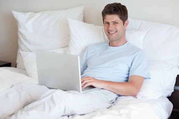 Улыбающийся мужчина лежит в постели с ноутбуком — стоковое фото